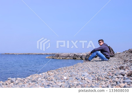 Young man traveler sits on stone beach and looks at sea on spring vacation. Young man traveler sits on stone beach and looks at sea on spring vacation. 133608030