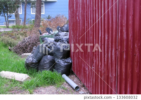 Black garbage bags near the fence of private house and an overturned trash can. 133608032
