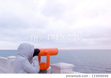 Woman in white jacket and hood looks in binoculars at sea waterfront with sea view. Side view. 133608046