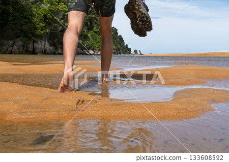 Hiker walking barefoot through water carrying shoes on Abel Tasman Track, NZ 133608592