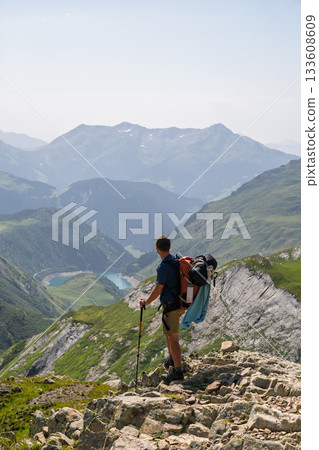 Hiker standing on ridge overlooking reservoir and valley in French Alps, TMB 133608609