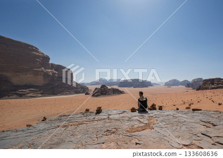 Tourist sitting on rocky ledge with stone cairns in Wadi Rum, Jordan Tourist sitting on rocky ledge with stone cairns in Wadi Rum, Jordan 133608636