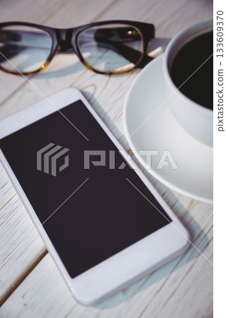 Showing pale wooden tabletop featuring white smartphone, black-framed eyeglasses and coffee cup 133609370