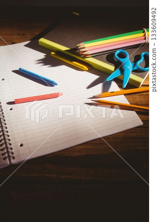 Spiral-bound notebook is lying open on dark wooden desk with crayons, pastel pencils and scissors Spiral-bound notebook is lying open on dark wooden desk with crayons, pastel pencils and scissors 133609492