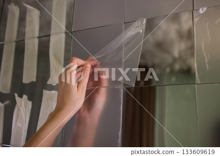 Removing protective layer to reveal clear glass, Carefully removing protective film from mirror to show pristine reflective surface in warm lit bathroom environment 133609619