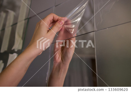 Removing protective layer to reveal clear glass, Carefully removing protective film from mirror to show pristine reflective surface in warm lit bathroom environment Removing protective layer to reveal clear glass, Carefully removing protective film from mirror to show pristine reflective surface in warm lit bathroom environment 133609620