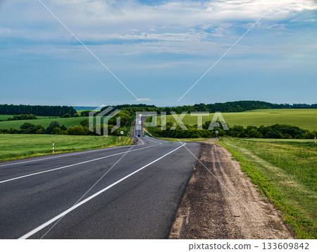 The side of the highway with a road running away into the distance between fields of wheat. Stormy skies. Trucks on a hilly road. 133609842