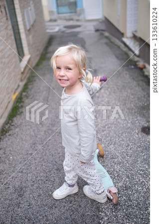 Little 4 Year Old Girl Walking Through Narrow Streets with a Scooter Without a Helmet 133610224