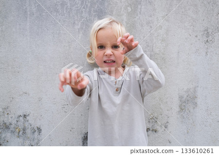 Little 4 Year Old Blonde Girl Standing by a Wall, Pretending to Be Angry and Showing Claws 133610261