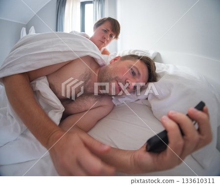 Wife curiously looking at her husband phone while he lies in bed.  133610311