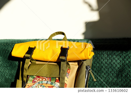 Stylish rolled-top yellow and green backpack with colorful patterned pocket on emerald velvet surface, sunlight. Tapestry and waterproof fabric 133610602