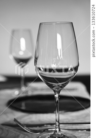 A refined black and white photograph of a table set for a formal meal, focusing on a clear wine glass in the foreground A refined black and white photograph of a table set for a formal meal, focusing on a clear wine glass in the foreground 133610724