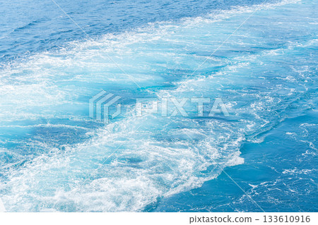 A bright view of a boat wake forming white foam patterns across the deep blue water. The dynamic movement and vivid turquoise shades create a refreshing, energetic seaside atmosphere. A bright view of a boat wake forming white foam patterns across the deep blue water. The dynamic movement and vivid turquoise shades create a refreshing, energetic seaside atmosphere. 133610916