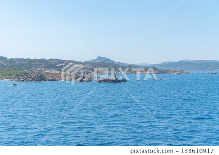 Calm blue water stretches toward a rocky shoreline with a solitary lighthouse standing on the stone edge. The landscape conveys a peaceful maritime atmosphere and a sense of open coastal space. 133610917