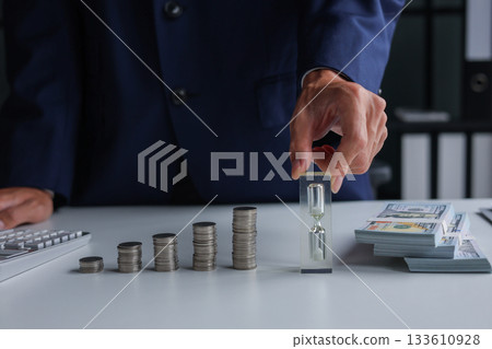 The concept of time and money. Hand holding a small hourglass next to stacks of coins and cash. 133610928