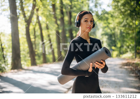 Model is with yoga mat. Young sportive skinny woman is in black fitness clothes outdoors 133611212