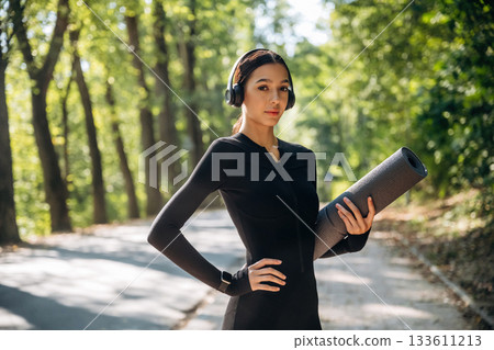 Skinny body type. Standing with yoga mat. Young sportive woman is in black fitness clothes outdoors 133611213