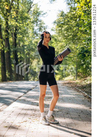 Skinny body type. Standing with yoga mat. Young sportive woman is in black fitness clothes outdoors 133611214
