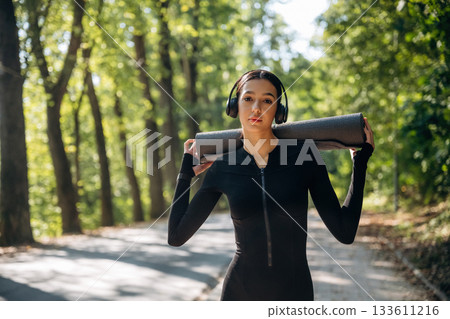 Skinny body type. Standing with yoga mat. Young sportive woman is in black fitness clothes outdoors Skinny body type. Standing with yoga mat. Young sportive woman is in black fitness clothes outdoors 133611216