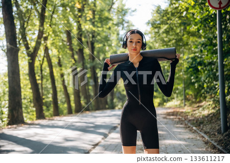 Skinny body type. Standing with yoga mat. Young sportive woman is in black fitness clothes outdoors Skinny body type. Standing with yoga mat. Young sportive woman is in black fitness clothes outdoors 133611217