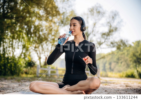 Rural place. Drinking water. Sitting. Young sportive skinny woman is in black fitness clothes outdoors 133611244