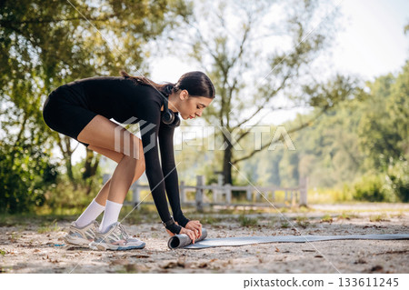 Rolling off the yoga mat on the ground. Young sportive skinny woman is in black fitness clothes outdoors 133611245