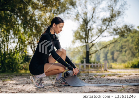Rolling off the yoga mat on the ground. Young sportive skinny woman is in black fitness clothes outdoors 133611246