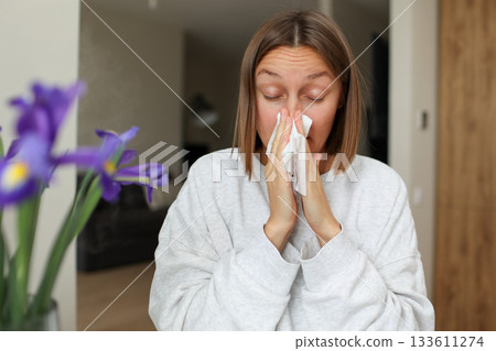 Seasonal allergy. Close up face of allergic young woman uses paper tissues from runny nose, sneezes from flowers pollen at home. Girl with close eyes with flu, itching or cough, allergy rhinitis Seasonal allergy. Close up face of allergic young woman uses paper tissues from runny nose, sneezes from flowers pollen at home. Girl with close eyes with flu, itching or cough, allergy rhinitis 133611274