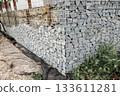 Stone fence in steel mesh and green lawn. Close up of gabion with tightly packed stones of varying sizes and textures. Combination of natural materials and industrial design. 133611281