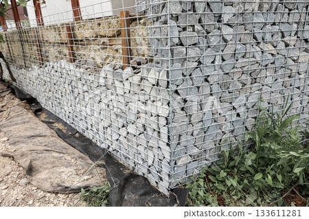 Stone fence in steel mesh and green lawn. Close up of gabion with tightly packed stones of varying sizes and textures. Combination of natural materials and industrial design. Stone fence in steel mesh and green lawn. Close up of gabion with tightly packed stones of varying sizes and textures. Combination of natural materials and industrial design. 133611281
