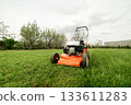Gardening work tools. Wide angle view of orange electric lawn mower, wheels, motor on bright lush green grass at backyard. Rotary lawn mower machine cut lawn. Professional lawn care service. 133611283