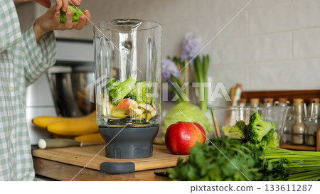 Unknown woman makes healthy detox breakfast smoothie at home. Female hands throws parsley in blender bowl, prepares fresh green vegetarian fruit-greens-vegetables drink to cleanse, provide energy body Unknown woman makes healthy detox breakfast smoothie at home. Female hands throws parsley in blender bowl, prepares fresh green vegetarian fruit-greens-vegetables drink to cleanse, provide energy body 133611287