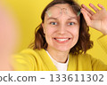 Playful young woman takes a selfie, smiles, sticks out her tongue, holds glasses on yellow backdrop. Modern lifestyle, social media, youth influencer marketing, self expression, personal branding 133611302