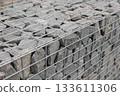 Stone fence in steel mesh and green lawn. Close up of gabion with tightly packed stones of varying sizes and textures. Combination of natural materials and industrial design. 133611306