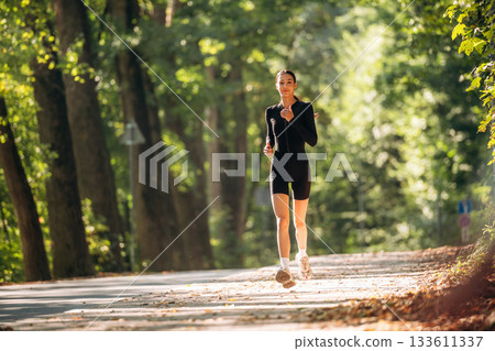 Jogging, front view. Young sportive skinny woman is in black fitness clothes outdoors Jogging, front view. Young sportive skinny woman is in black fitness clothes outdoors 133611337
