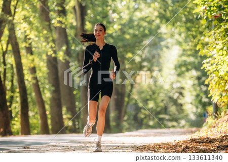 Jogging, front view. Young sportive skinny woman is in black fitness clothes outdoors Jogging, front view. Young sportive skinny woman is in black fitness clothes outdoors 133611340