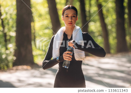 With bottle of water. Young sportive skinny woman is in black fitness clothes outdoors With bottle of water. Young sportive skinny woman is in black fitness clothes outdoors 133611343