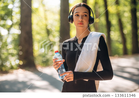 With bottle of water. Young sportive skinny woman is in black fitness clothes outdoors With bottle of water. Young sportive skinny woman is in black fitness clothes outdoors 133611346