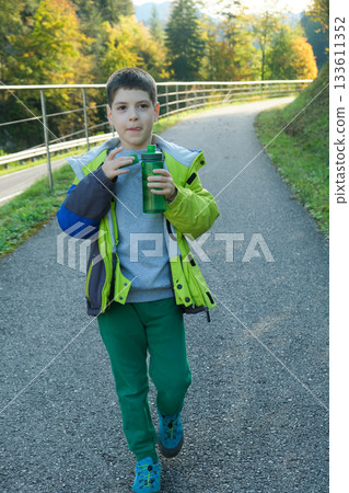 A schoolchild walks around the nature and drinks water from a plastic bottle. 133611352