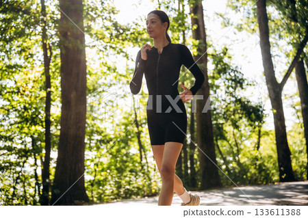 Running, jogging in the forest. Young sportive skinny woman is in black fitness clothes outdoors Running, jogging in the forest. Young sportive skinny woman is in black fitness clothes outdoors 133611388