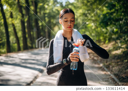 White towel is on the neck. Young sportive skinny woman is in black fitness clothes outdoors White towel is on the neck. Young sportive skinny woman is in black fitness clothes outdoors 133611414