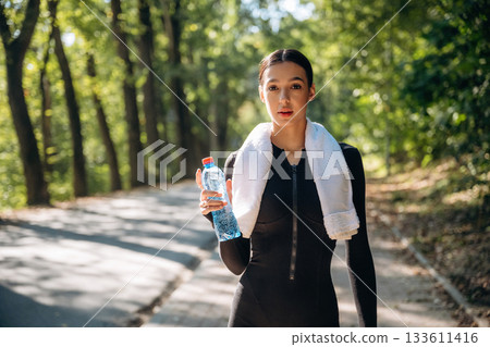 Stay hydrated, with bottle of water, standing. Young sportive skinny woman is in black fitness clothes outdoors Stay hydrated, with bottle of water, standing. Young sportive skinny woman is in black fitness clothes outdoors 133611416