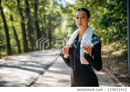Stay hydrated, with bottle of water, standing. Young sportive skinny woman is in black fitness clothes outdoors Stay hydrated, with bottle of water, standing. Young sportive skinny woman is in black fitness clothes outdoors 133611422