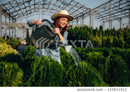 Standing and watering the plants. Young redhead woman is taking care of garden Standing and watering the plants. Young redhead woman is taking care of garden 133611912