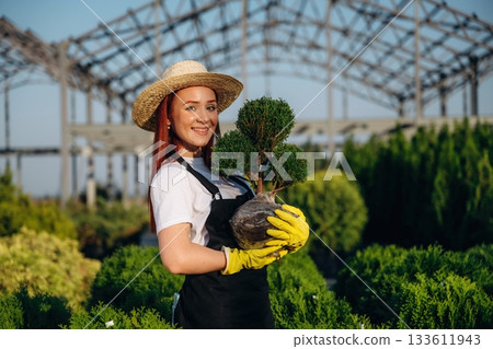Cheerful facial expression, holding plant. Young redhead woman is taking care of garden 133611943