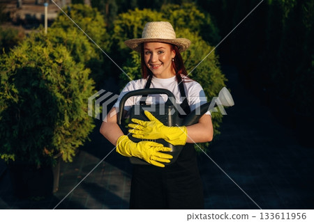 Black watering can made of plastic. Young redhead woman is taking care of garden Black watering can made of plastic. Young redhead woman is taking care of garden 133611956
