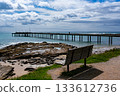 A wooden bench on the shore and pier extended to the blue sea. 133612736