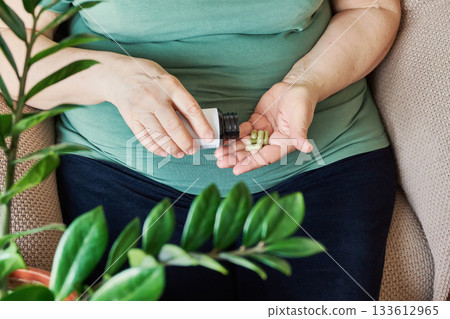 Mature woman with overweight pouring green capsules from bottle into her hand for daily health Mature woman with overweight pouring green capsules from bottle into her hand for daily health 133612965