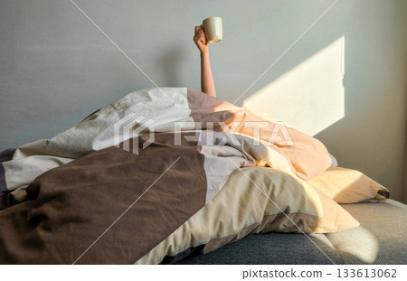 A woman's hand with a cup of coffee in bed in the light of the morning sun's rays. Good sunny cheerful morning. A woman's hand with a cup of coffee in bed in the light of the morning sun's rays. Good sunny cheerful morning. 133613062