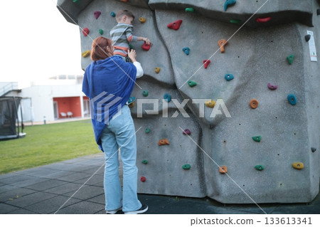 Mother assisting boy climbing outdoor rock wall 133613341